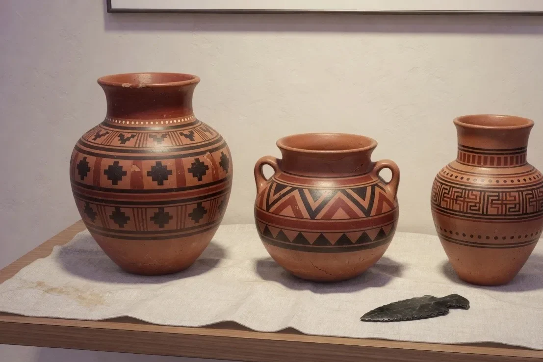 Três vasilhas de barro e uma lâmina de pedra sobre uma mesa clara, sem ficha ao lado.