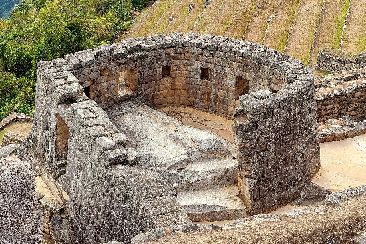 Muro de pedra curvo do Templo do Sol, com janelas trapezoidais talhadas na rocha e terraços verdes ao fundo, sob luz do dia.