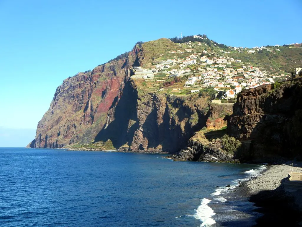 O paredão do Cabo Girão, uma falésia alta de rocha avermelhada que cai direto no mar azul, com casas no topo verde.