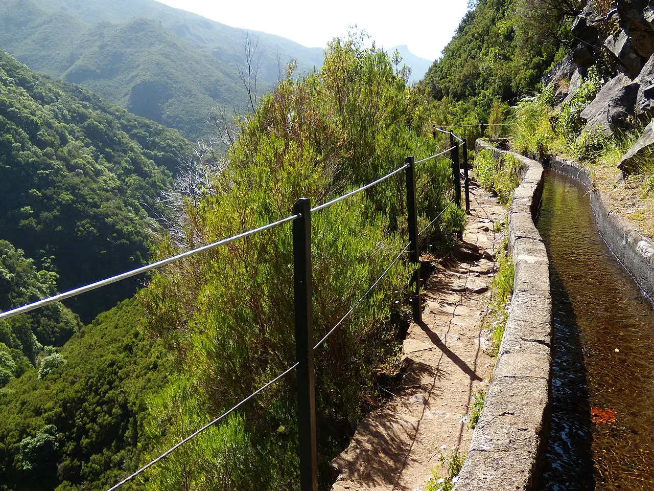 A trilha da Levada das 25 Fontes ao lado do canal de água, protegida por uma cerca de arame, numa encosta verde e íngreme.