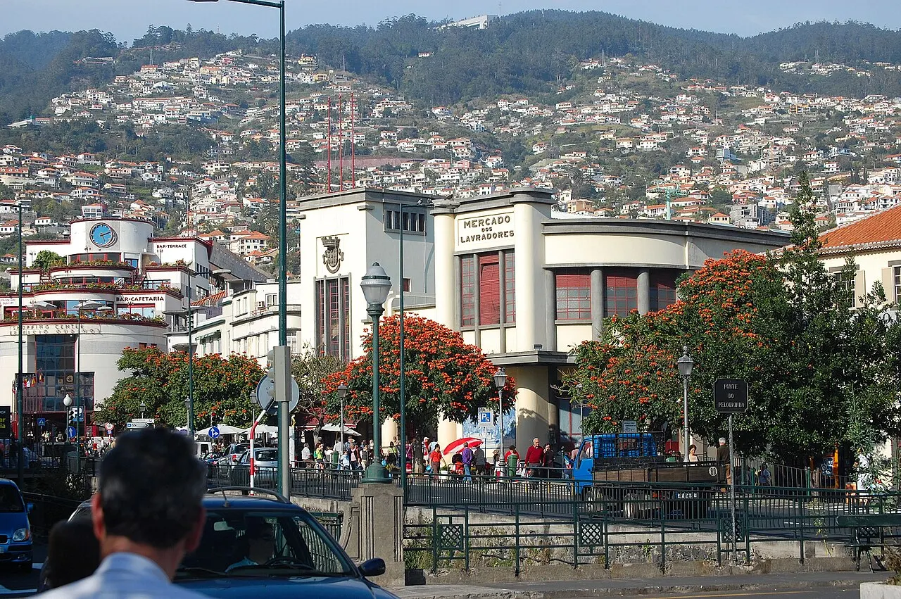 O prédio art déco do Mercado dos Lavradores numa esquina do Funchal, com árvores floridas à frente e a encosta cheia de casas atrás.