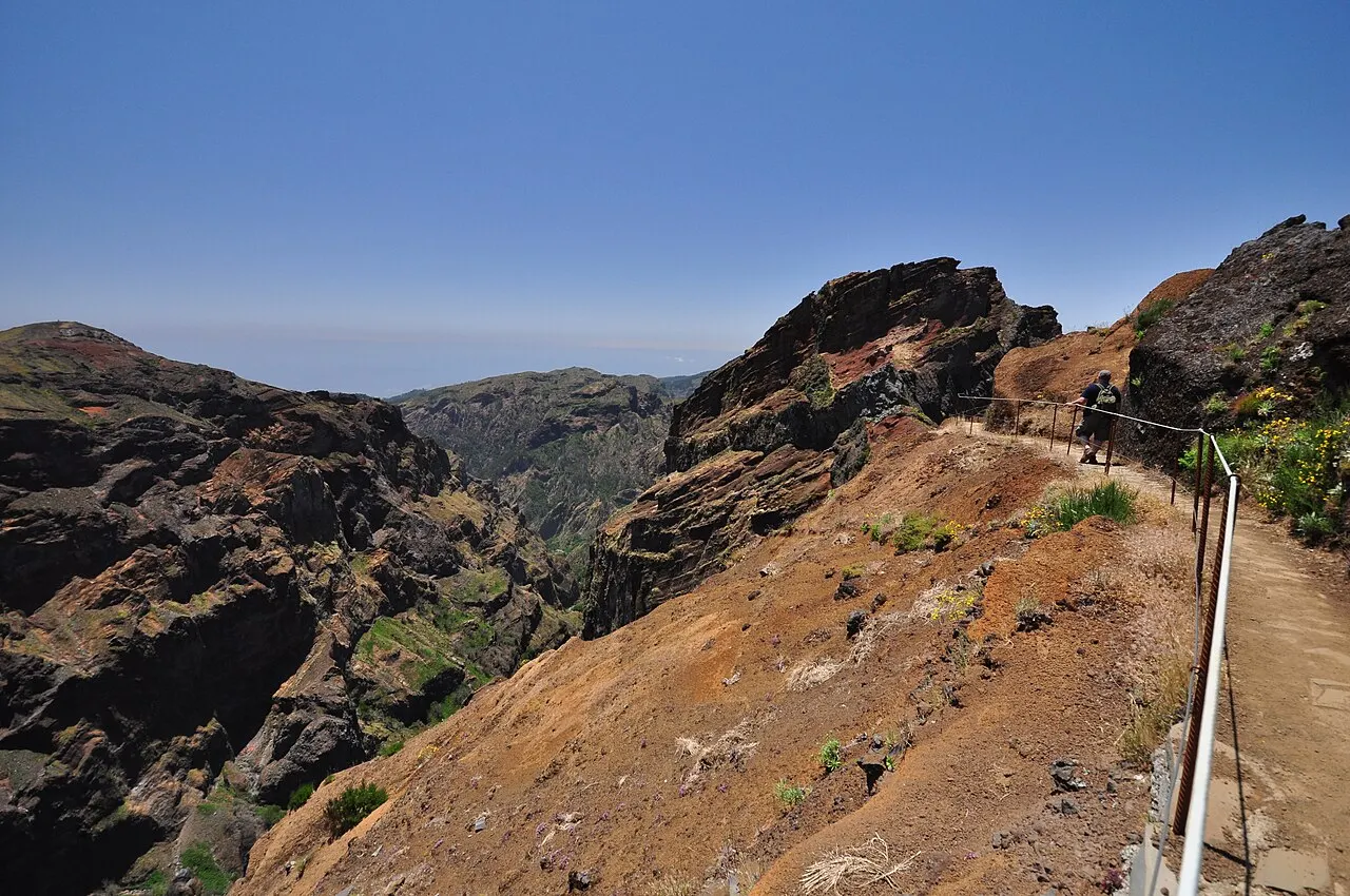 Uma trilha estreita com corrimão seguindo uma crista de terra vermelha entre picos de rocha escura, perto do Pico do Arieiro, sob céu azul.