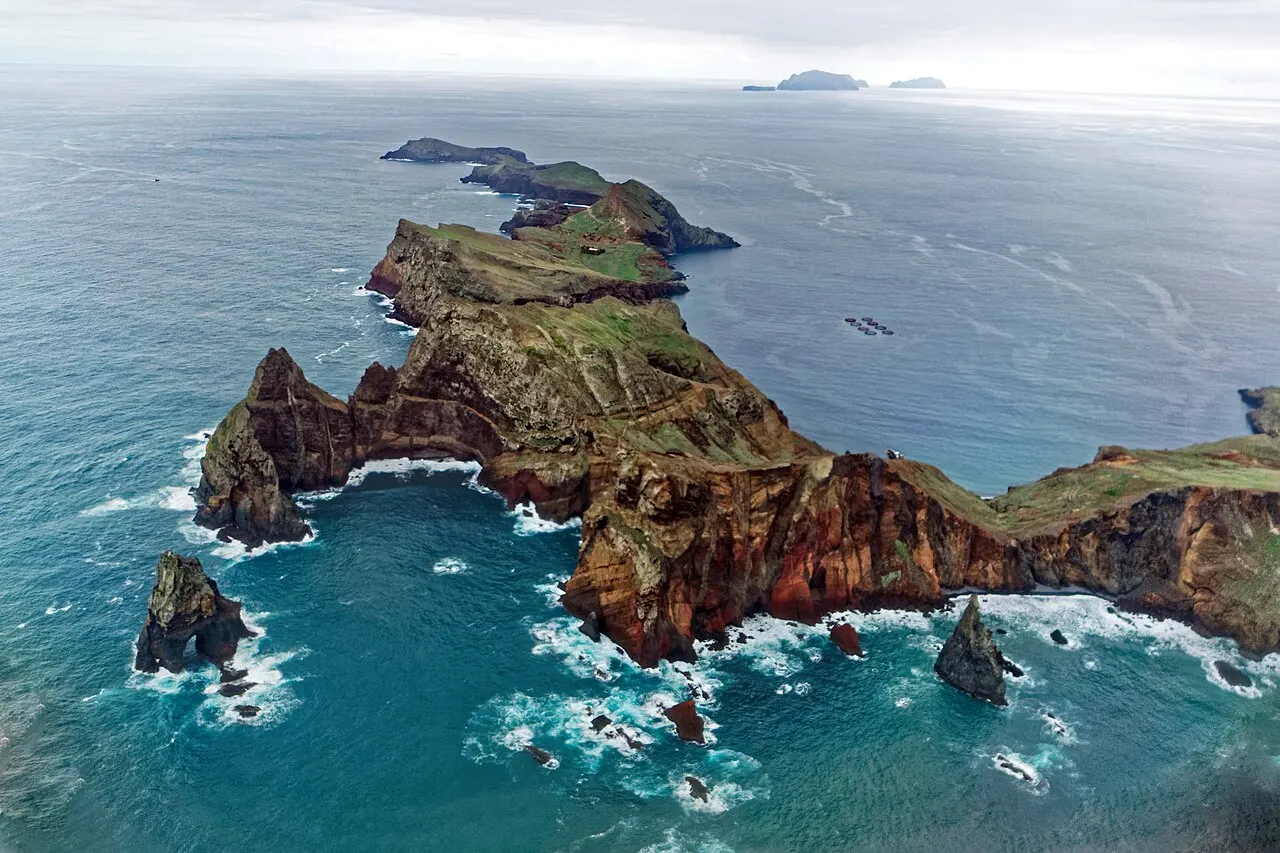 Vista aérea da Ponta de São Lourenço: a península estreita de rocha avermelhada e capim verde entrando no mar, com ilhéus na ponta.