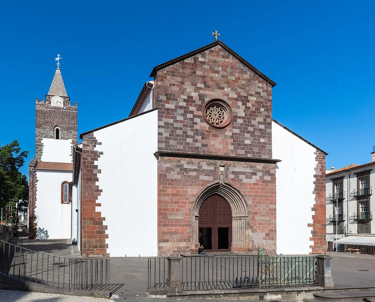 A Sé do Funchal: fachada de pedra vulcânica escura com rosácea, paredes caiadas de branco e a torre do sino à esquerda, sob céu azul.