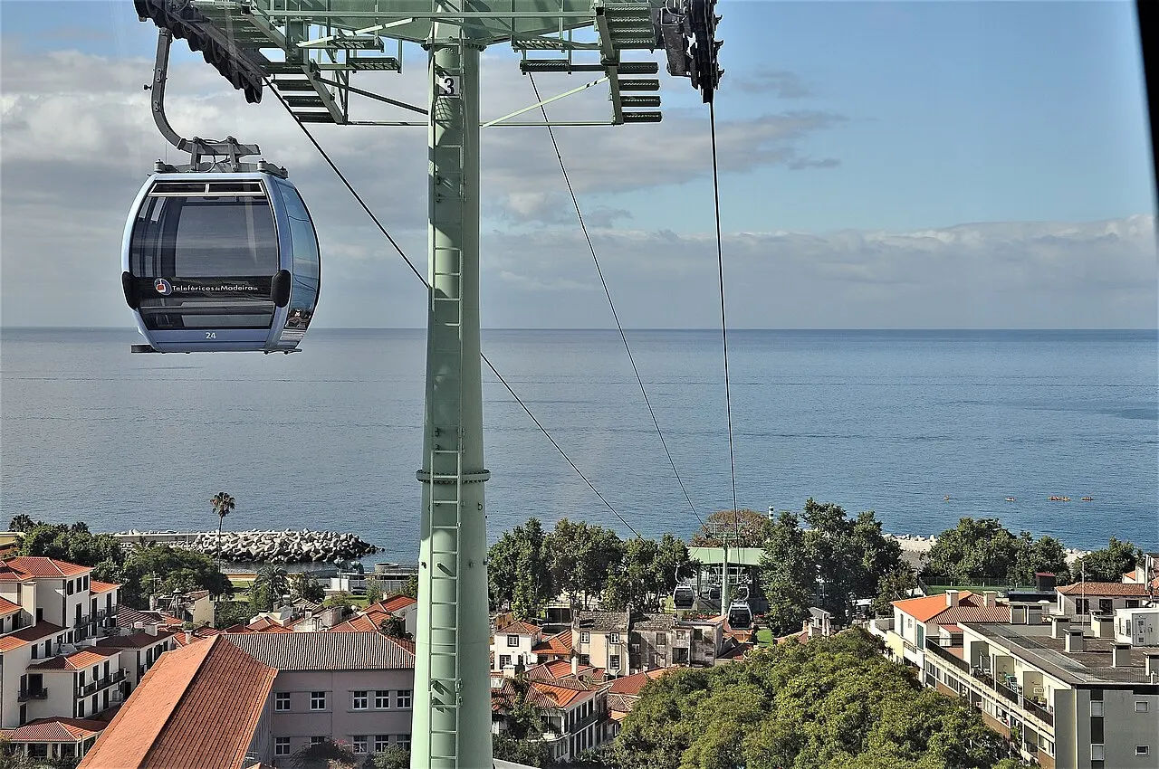 Uma cabine do teleférico do Funchal pendurada no cabo, com um pilar verde, os telhados da cidade embaixo e o mar ao fundo.