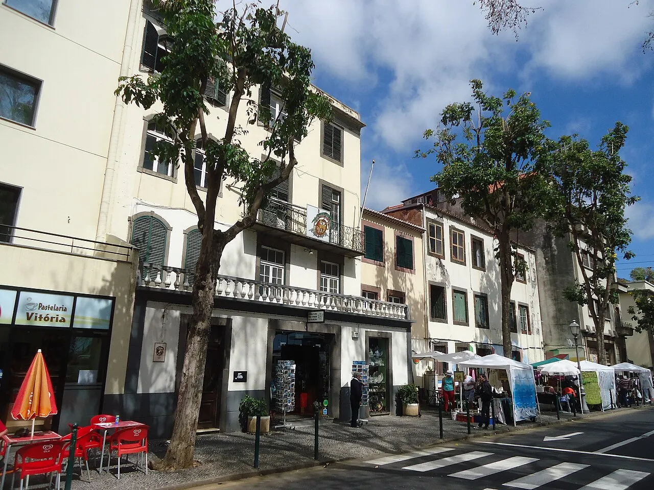 Uma rua da Zona Velha do Funchal com casas antigas de varanda, árvores na calçada e mesas de bar com guarda-sóis vermelhos.