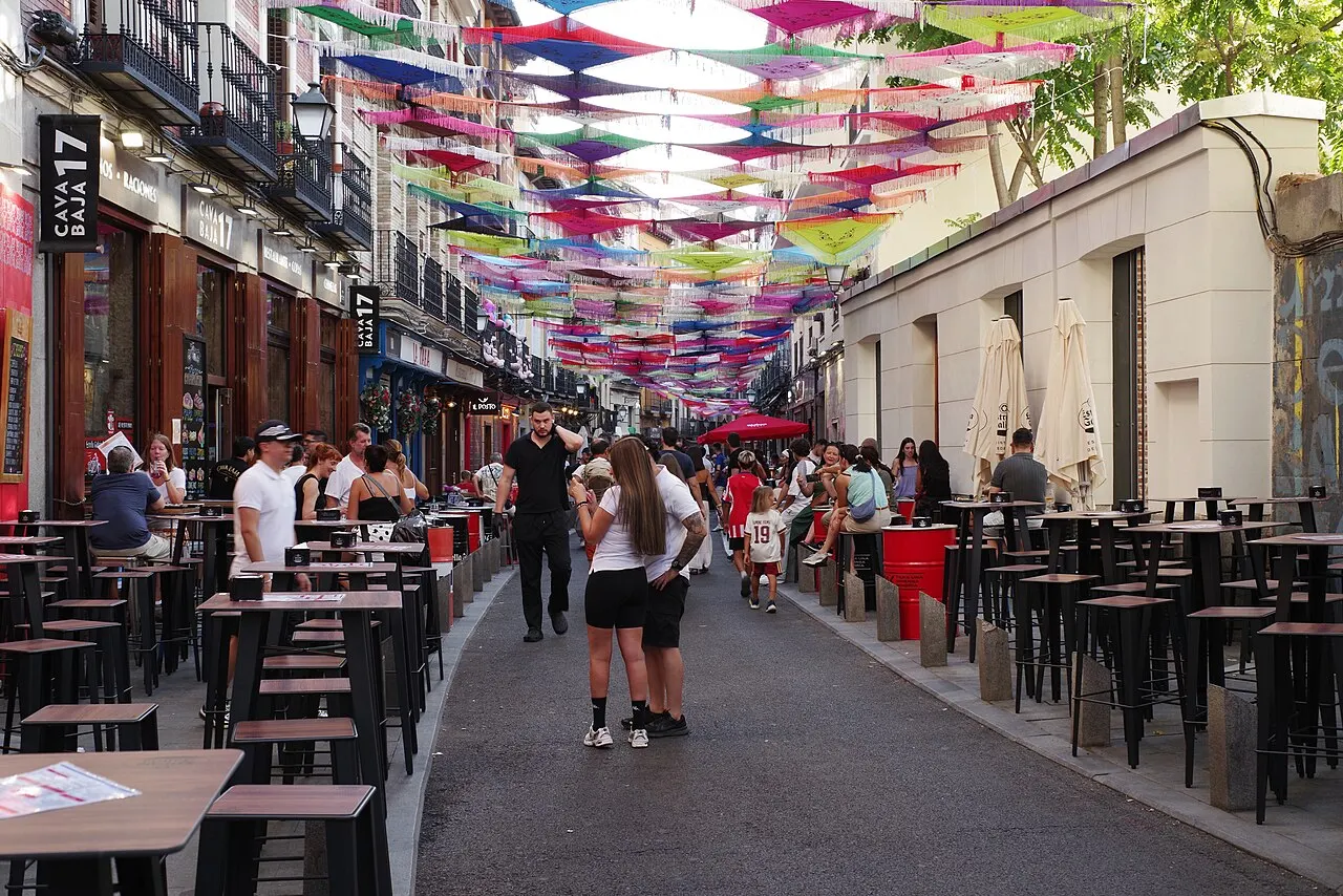 Rua estreita coberta por fileiras de guarda-sóis coloridos suspensos, com mesas altas e bancos de bares nas calçadas cheios de gente em dia de sol.