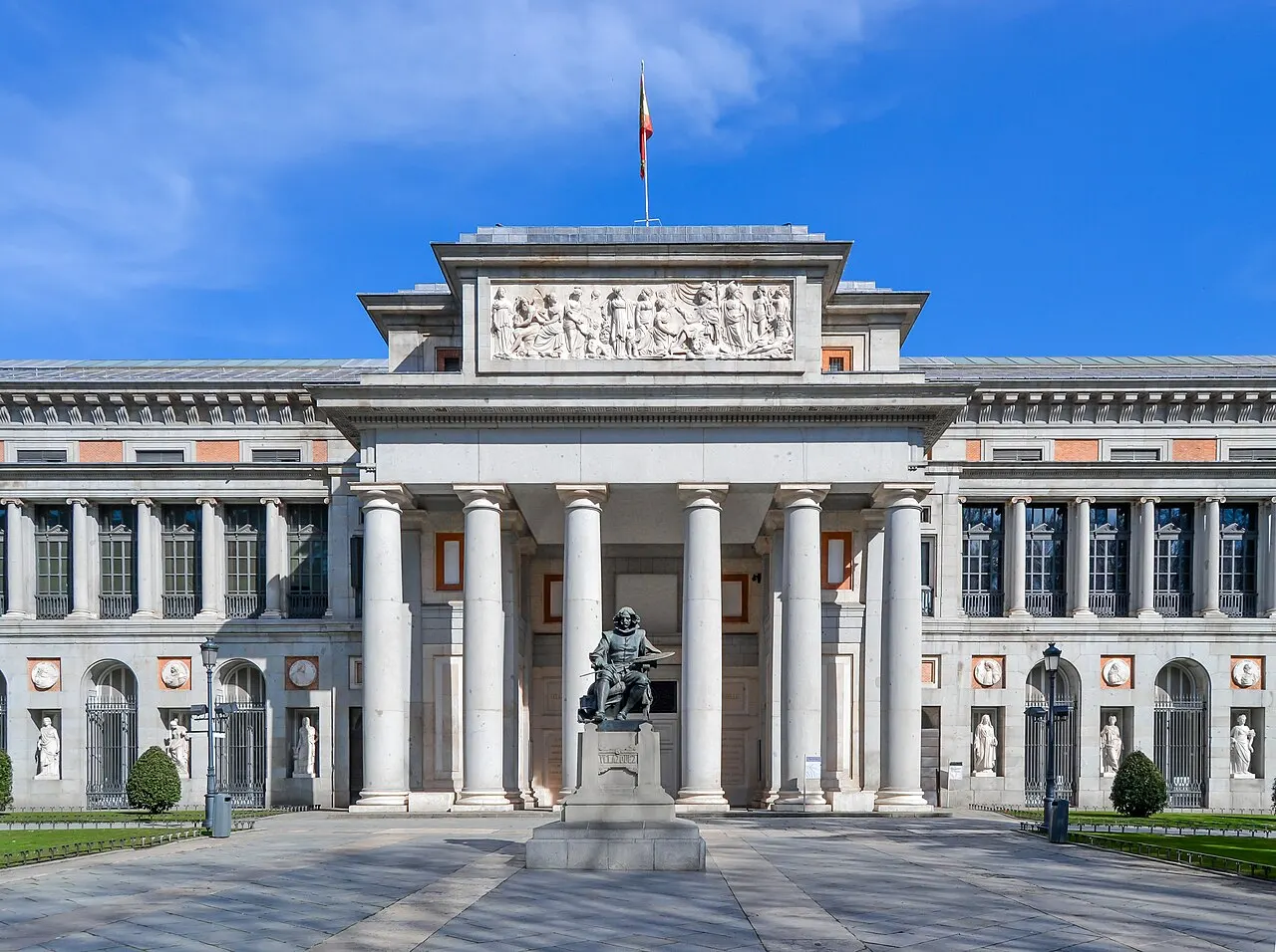 Fachada neoclássica do Museu do Prado, com pórtico de colunas jônicas, friso esculpido no alto e a estátua sentada de Velázquez em primeiro plano, sob céu azul.