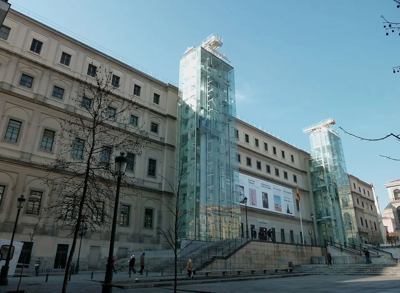 Fachada do Museu Reina Sofía, com torres de elevador em vidro anexadas ao edifício de pedra clara, pessoas caminhando na praça e céu azul ao fundo.