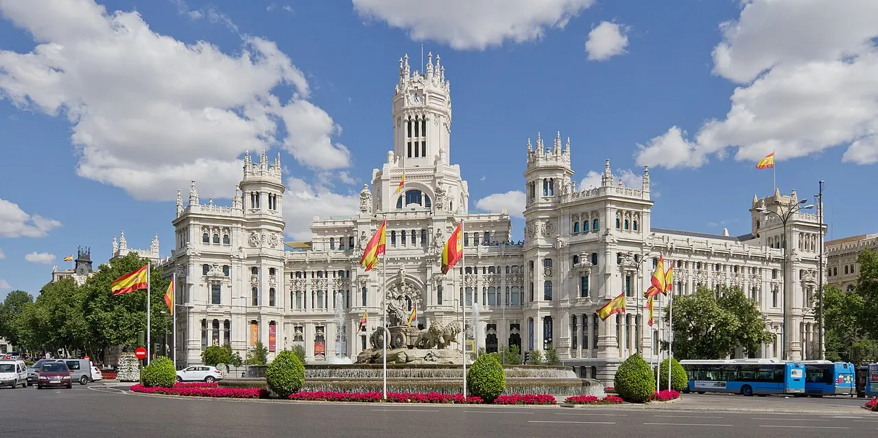 Fachada de pedra branca do Palacio de Cibeles, em Madri, com a torre central e as agulhas laterais, e árvores da praça cobrindo a base.