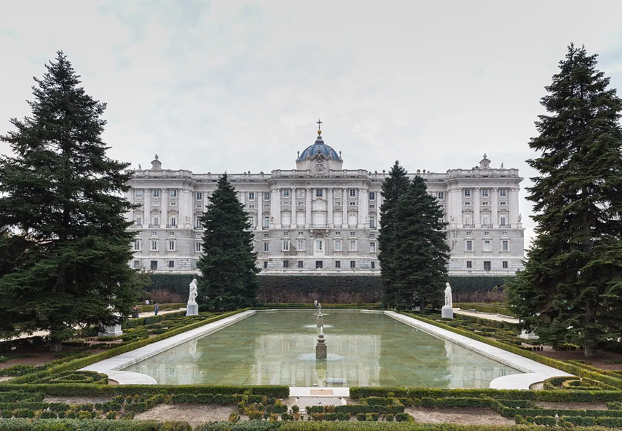 Fachada do Palácio Real de Madri, com a cúpula ao centro, vista de um jardim com espelho d'água e coníferas.