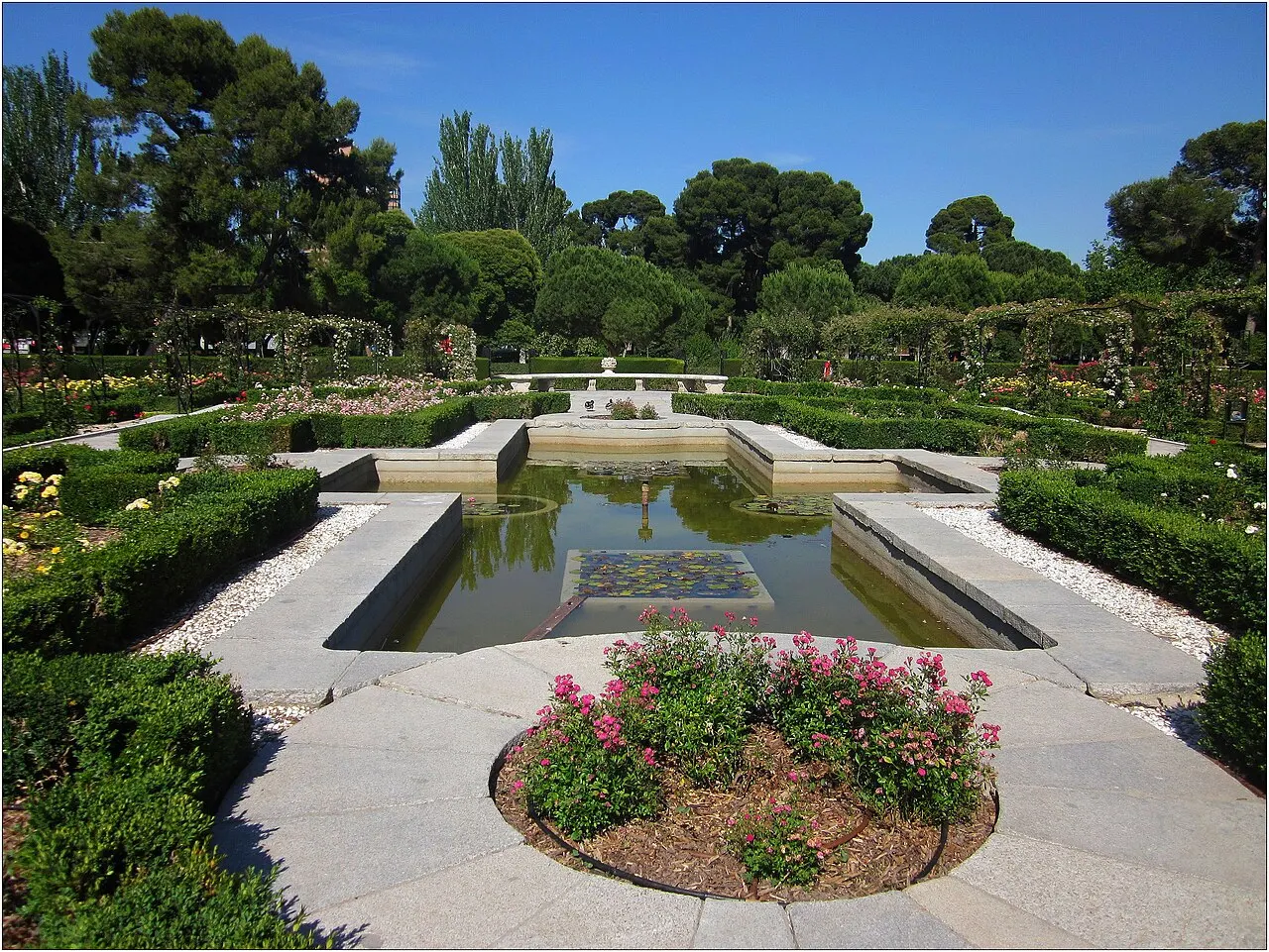 Jardim formal do Parque del Retiro, com espelho d'água retangular cercado de roseiras, arbustos aparados e árvores altas, sob céu azul claro.