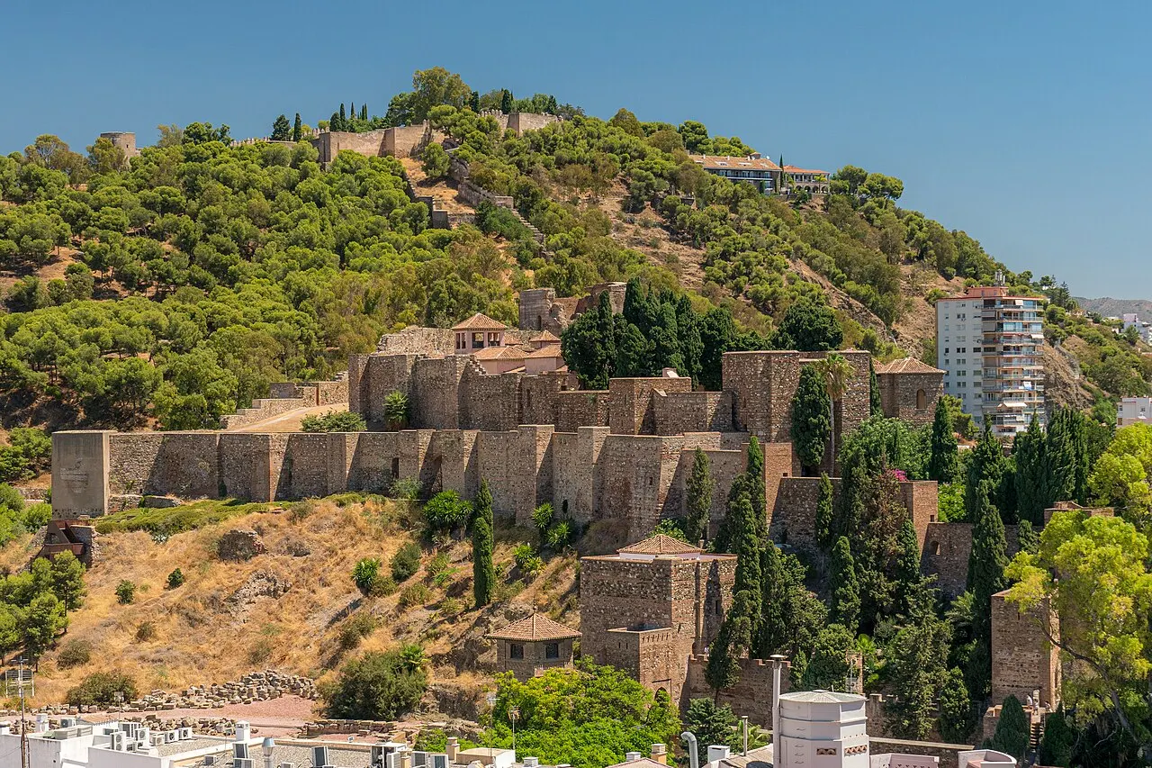 Les murailles et les tours de pierre de l'Alcazaba étagées sur la colline verte, avec le château de Gibralfaro au sommet.