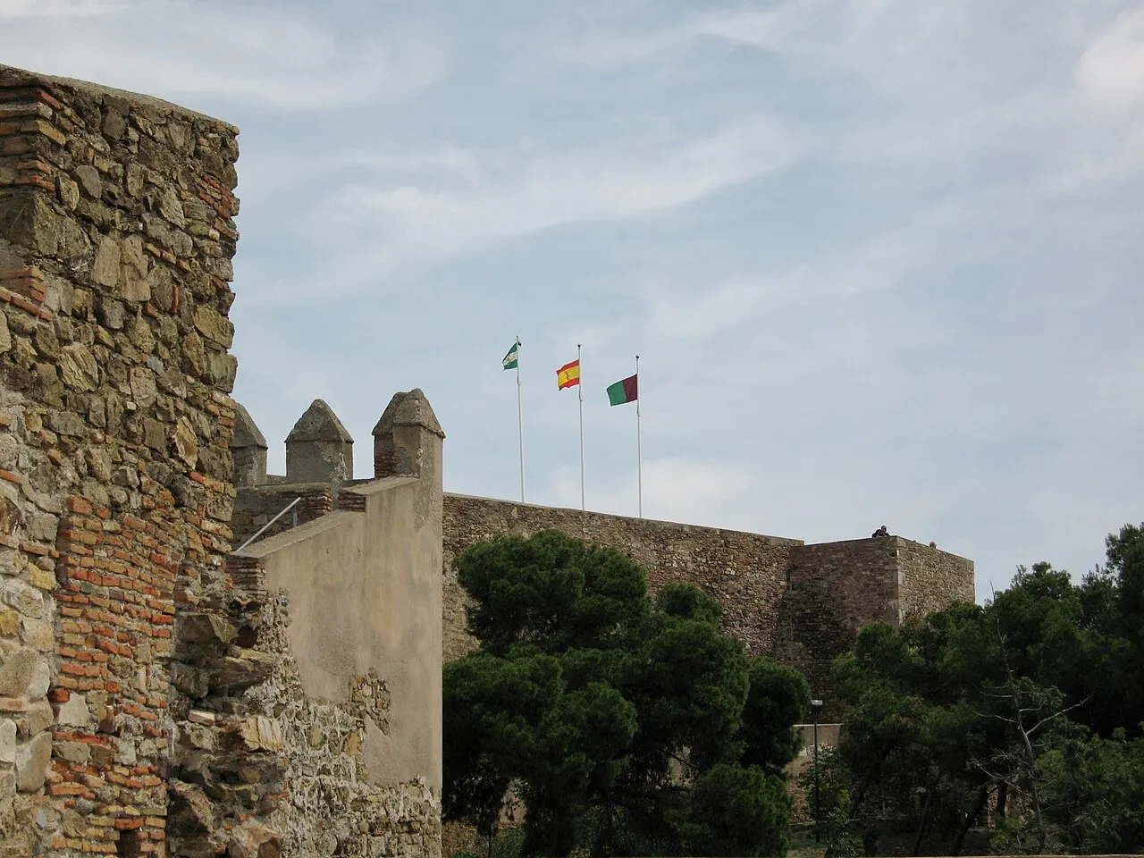 Les remparts de pierre du château de Gibralfaro avec créneaux et trois drapeaux au-dessus, parmi les pins.
