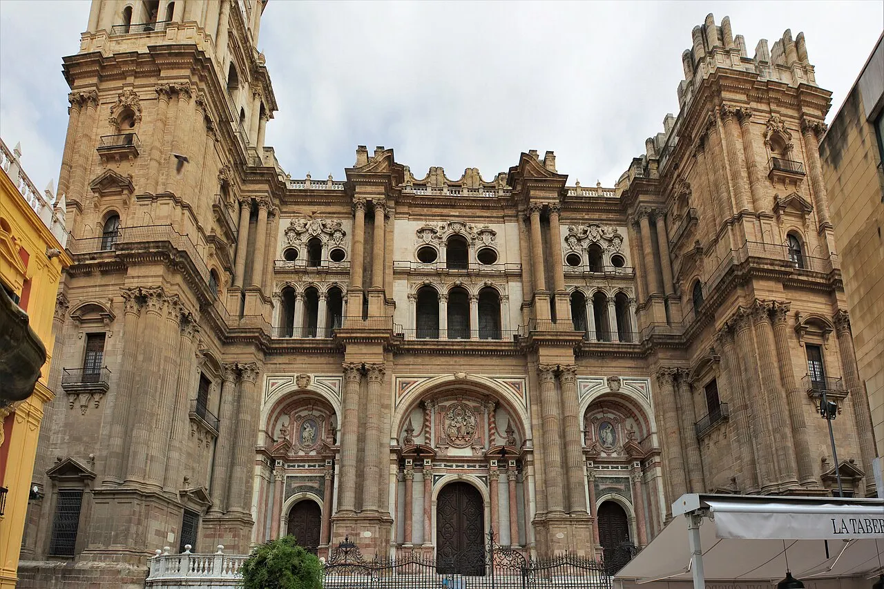 A fachada barroca da Catedral de Málaga, de pedra escura, com três portais, colunas e a torre à esquerda.