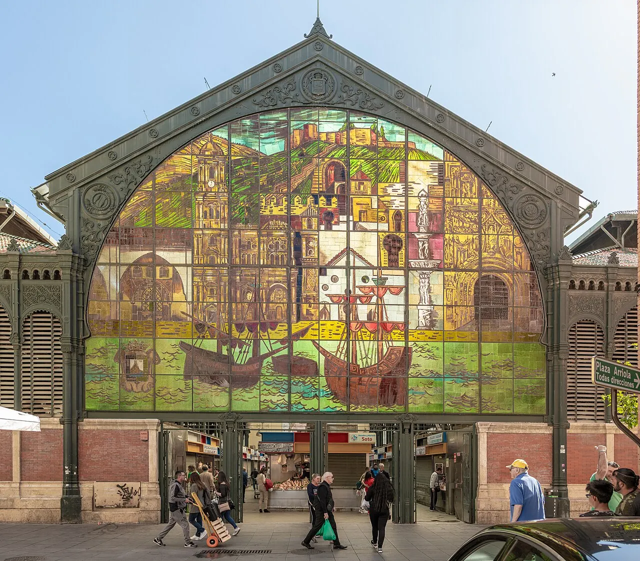 A fachada do Mercado de Atarazanas com o grande vitral colorido em arco sobre a entrada e pessoas passando embaixo.