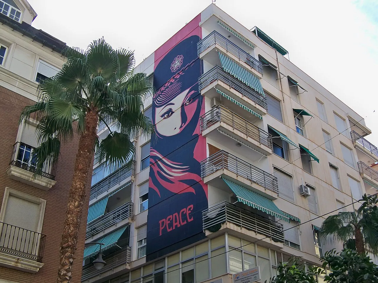 Um mural grande de Obey com uma mulher de lenço e a palavra PEACE pintado na lateral de um prédio do Soho, ao lado de uma palmeira.