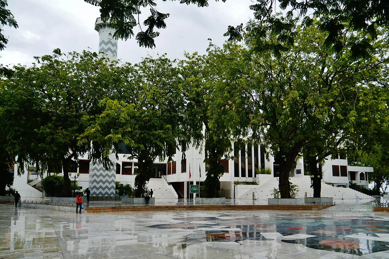 O prédio branco do Centro Islâmico de Malé atrás de árvores grandes, com o minarete à esquerda e a praça molhada de chuva à frente.