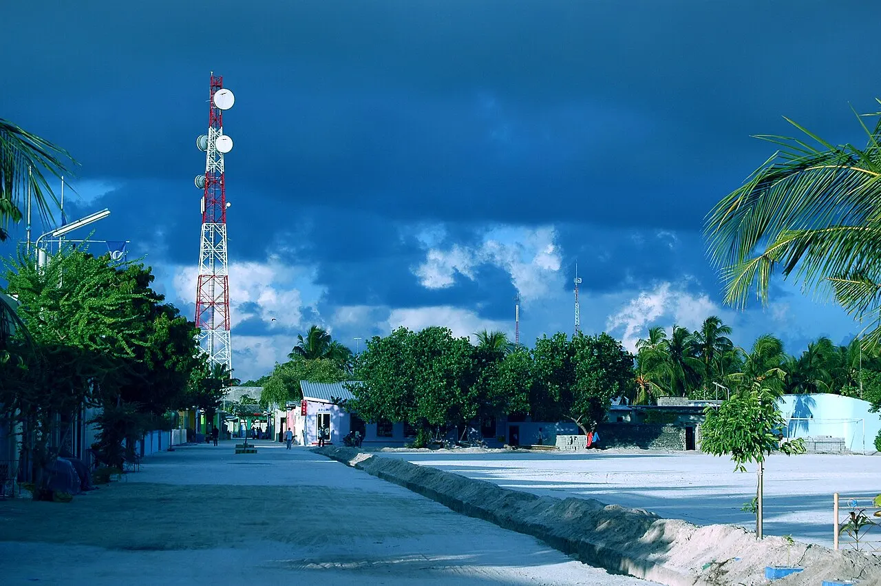 Uma rua larga de areia batida em Maafushi, com palmeiras, uma torre de antena e nuvens escuras de chuva no céu.