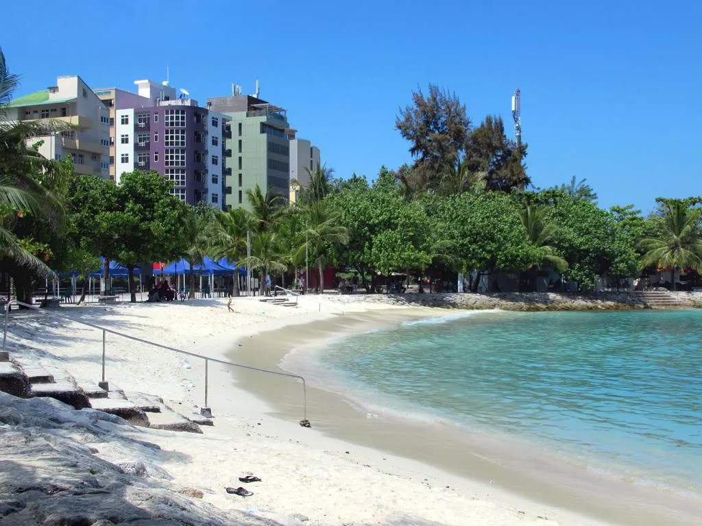 A Praia Artificial de Malé, uma curva curta de areia branca e água turquesa, com árvores e prédios altos logo atrás.