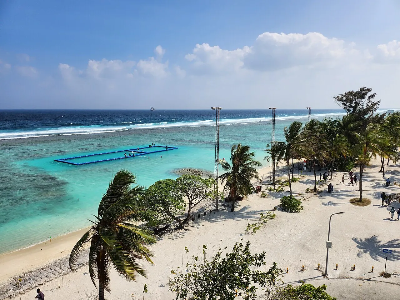 A praia de Hulhumalé vista do alto: areia branca com palmeiras, a lagoa turquesa e rasa e a linha do recife mais adiante.