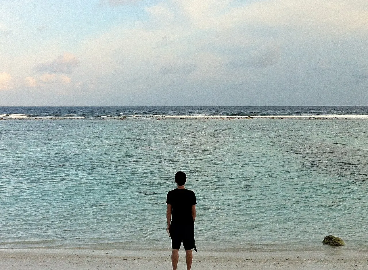 Uma pessoa de pé na água rasa e calma da beira de Villimalé, olhando o mar no fim de tarde, com o recife ao fundo.