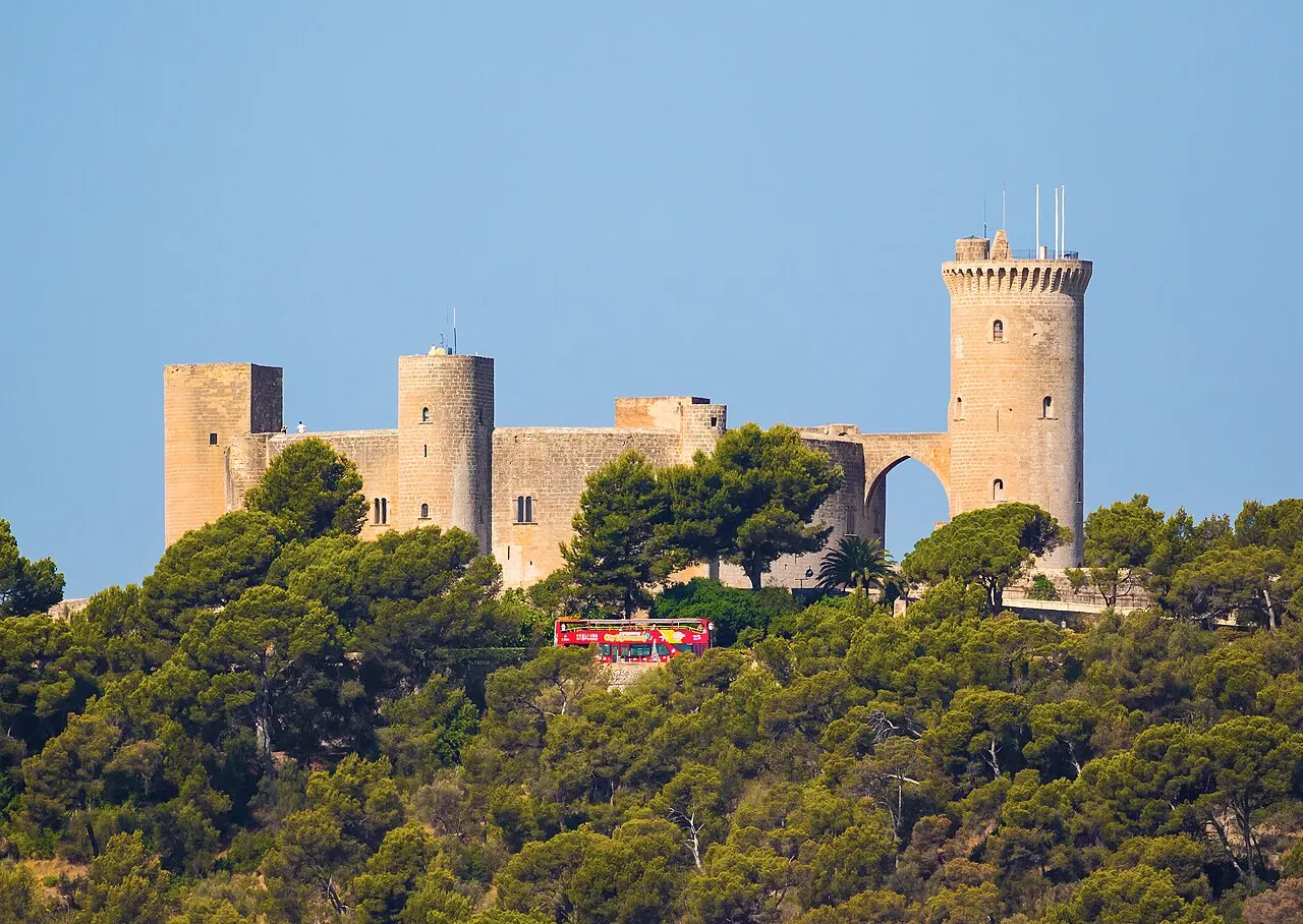 O Castelo de Bellver, de planta circular e torres redondas, no alto de um morro coberto de pinheiros, sob céu azul.