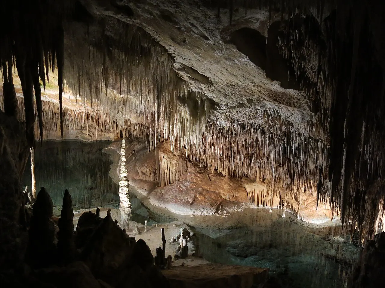 O interior das Cuevas del Drach, com estalactites claras penduradas do teto e um lago subterrâneo de água parada.