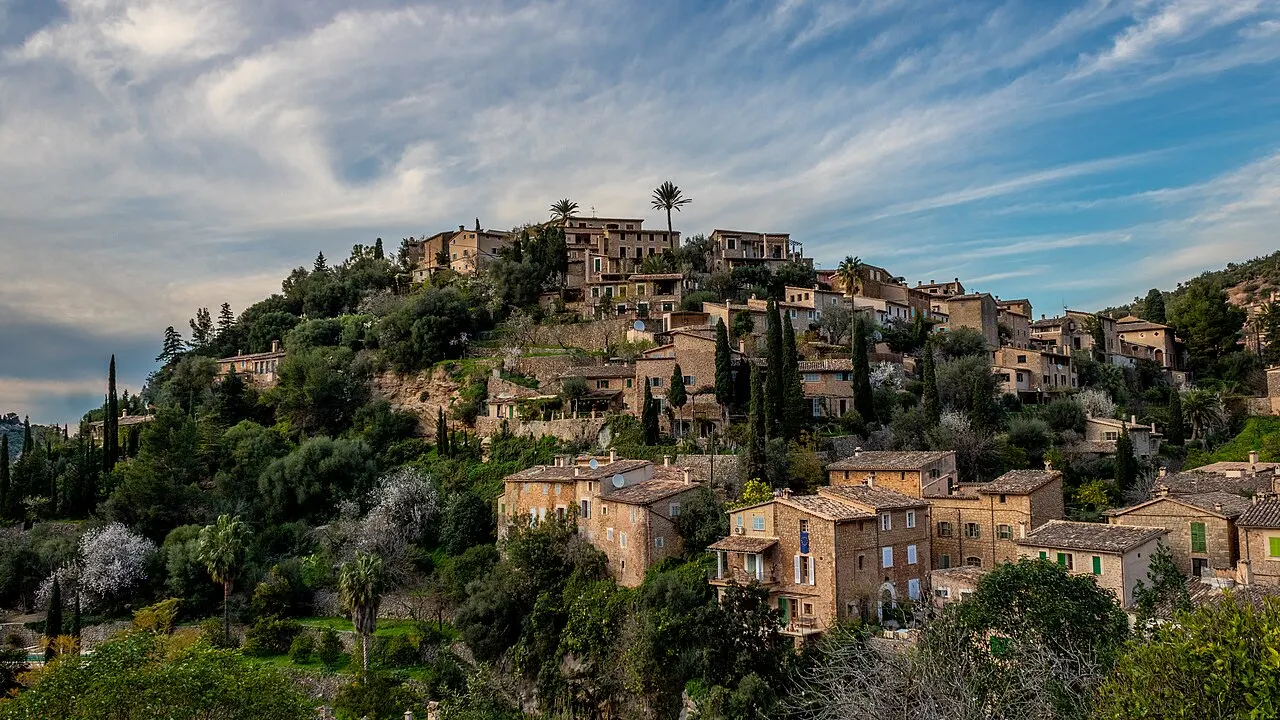 As casas de pedra de Deià agrupadas no alto de um morro verde, com a igreja no topo e nuvens no céu.