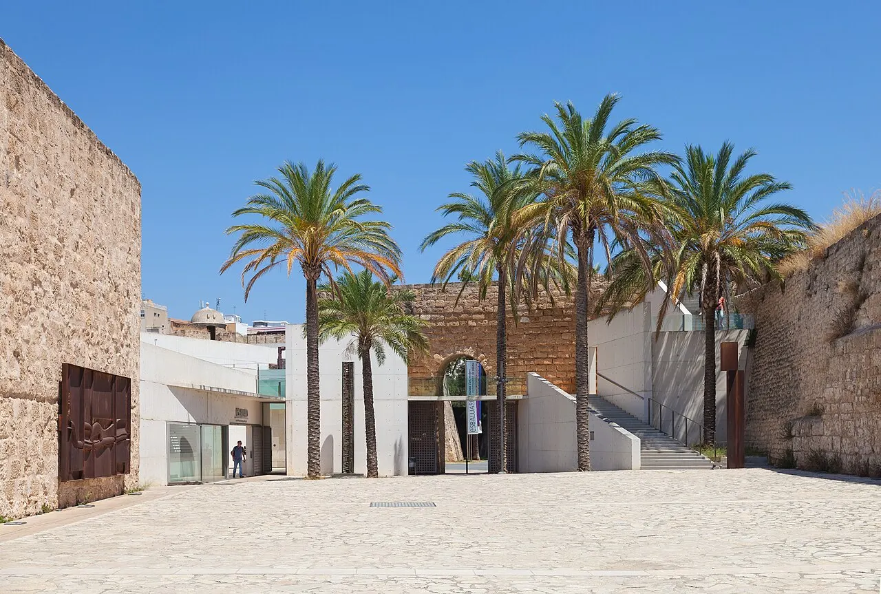 O pátio de entrada do Es Baluard entre as muralhas antigas de pedra, com o prédio branco do museu e palmeiras.