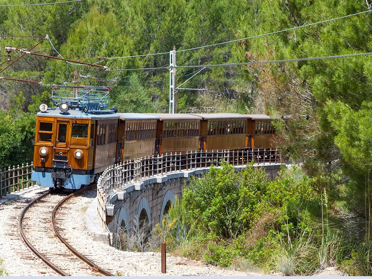 O trem antigo de madeira de Sóller fazendo uma curva num viaduto de pedra, entre pinheiros.