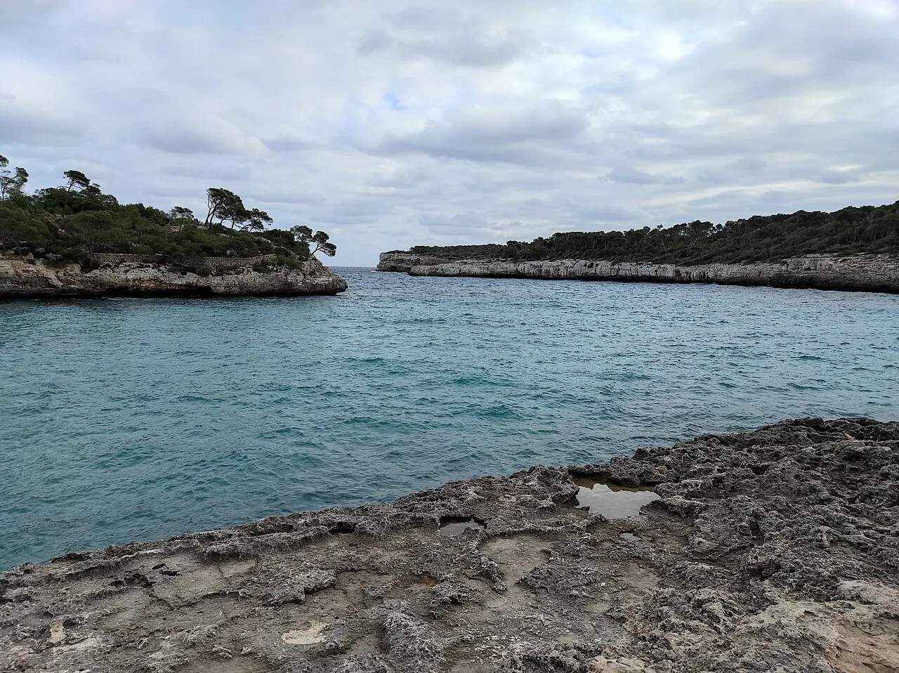 Uma enseada do Parque Natural de Mondragó, com água verde-azulada, rocha calcária em primeiro plano e pinheiros nas pontas.