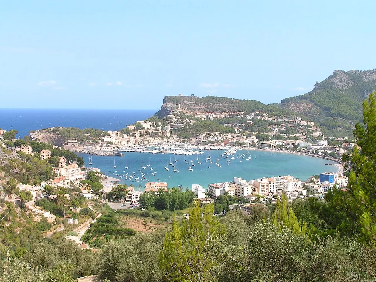 A baía em ferradura do Port de Sóller vista do alto, com barcos ancorados, casas na orla e o cabo rochoso atrás.