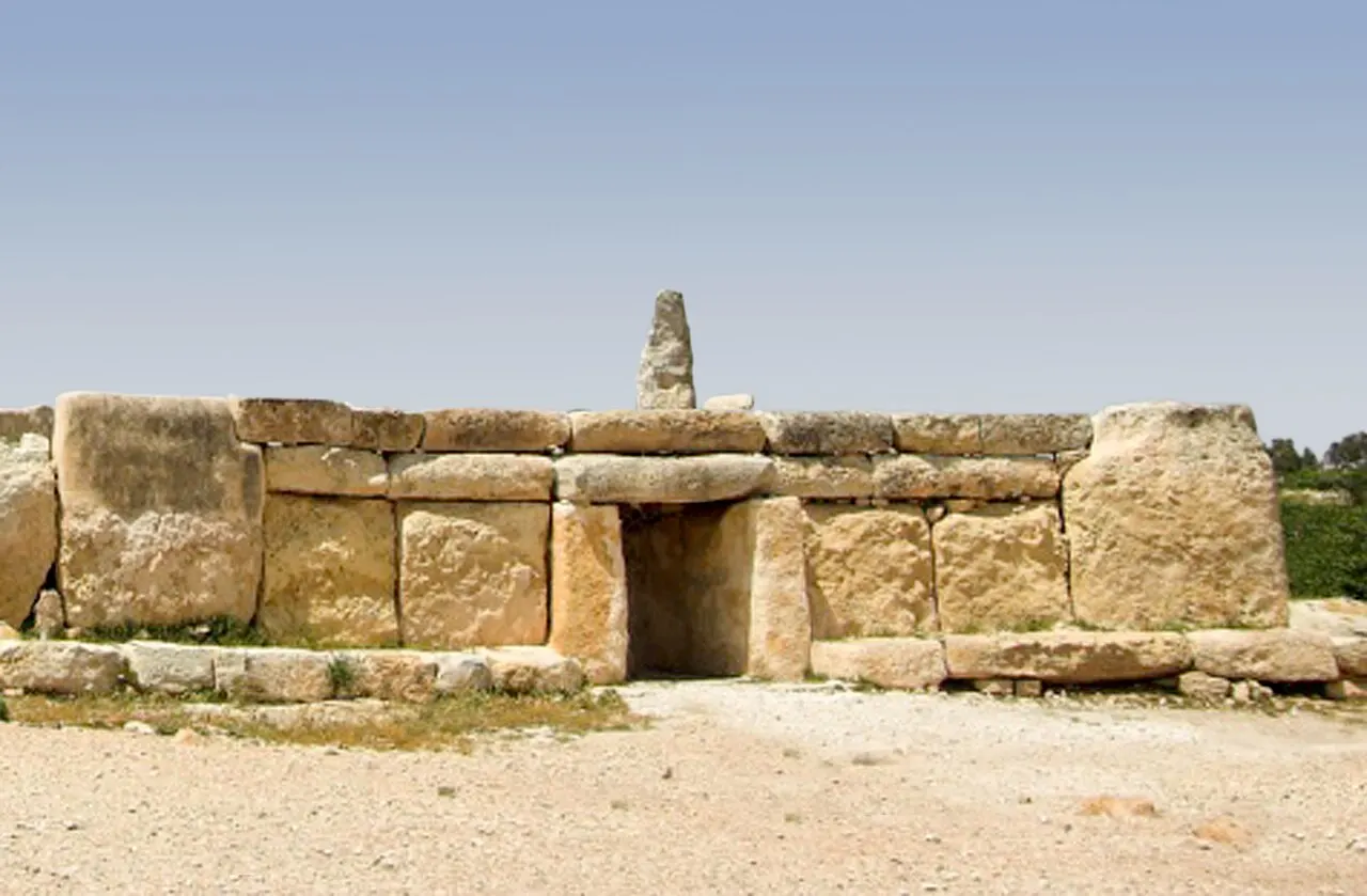 A fachada de grandes blocos de pedra do templo megalítico de Ħaġar Qim, com a porta trilítica no meio e um monólito atrás.