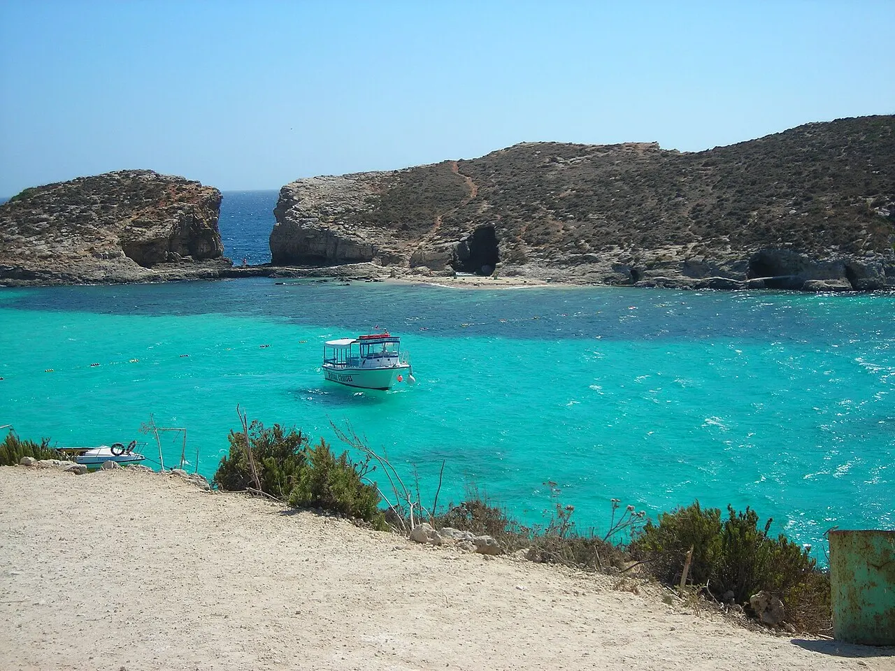 A água turquesa da Blue Lagoon de Comino com um barco ancorado, a ilhota de Cominotto atrás e o caminho de terra em primeiro plano.