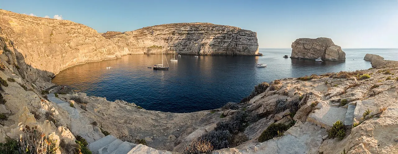 Panorama da baía de Dwejra, em Gozo, com a água escura, barcos ancorados, as falésias e a Fungus Rock à direita.