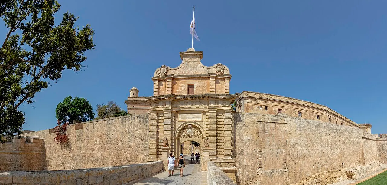 A Porta Principal barroca de Mdina no meio das muralhas de pedra, com a ponte de acesso e duas pessoas entrando.