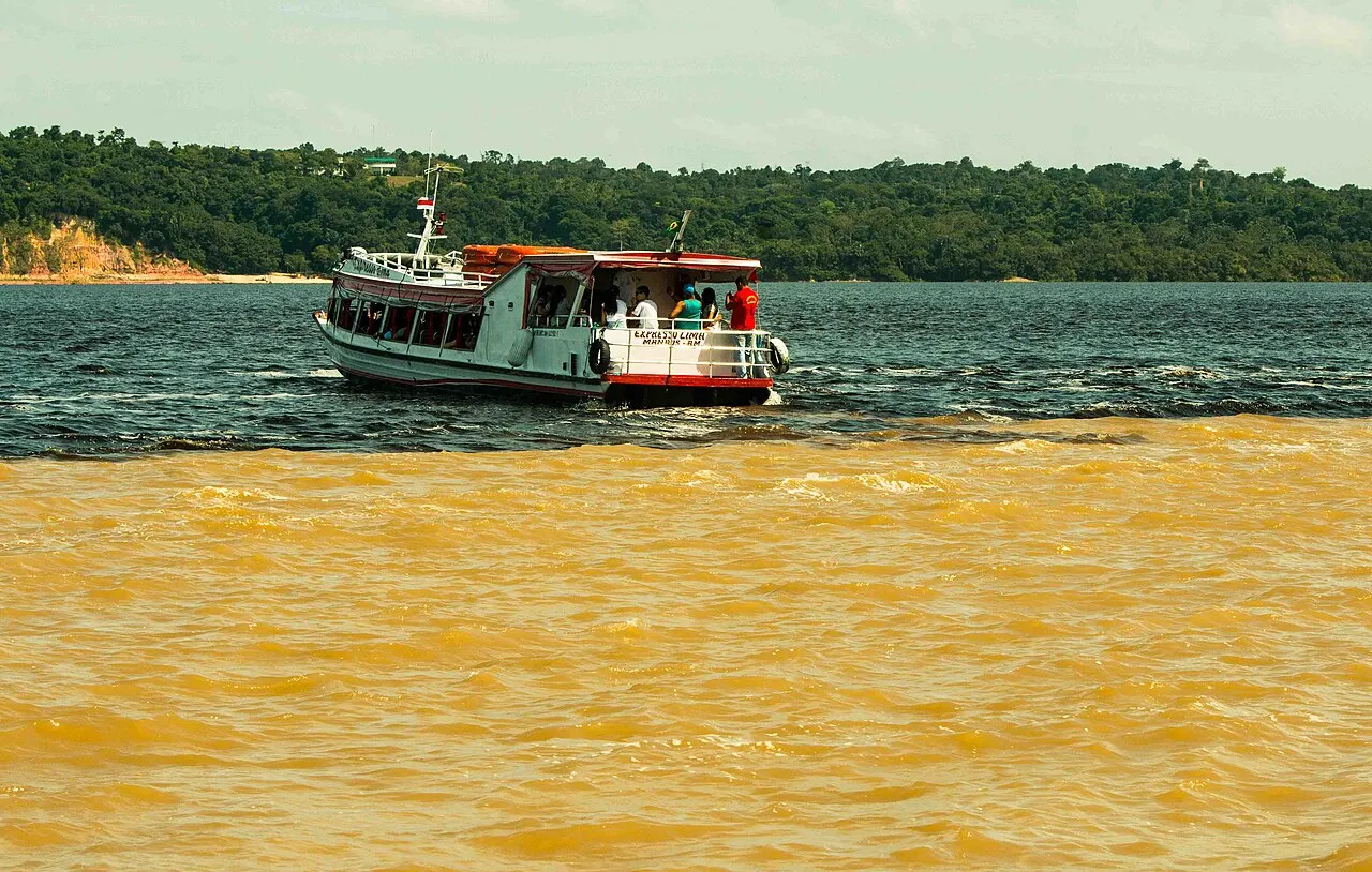 La línea nítida donde el agua oscura del río Negro se junta con el agua amarilla y turbia del Solimões, con un barco de pasajeros blanco y rojo navegando sobre ella y el monte bajo al fondo.