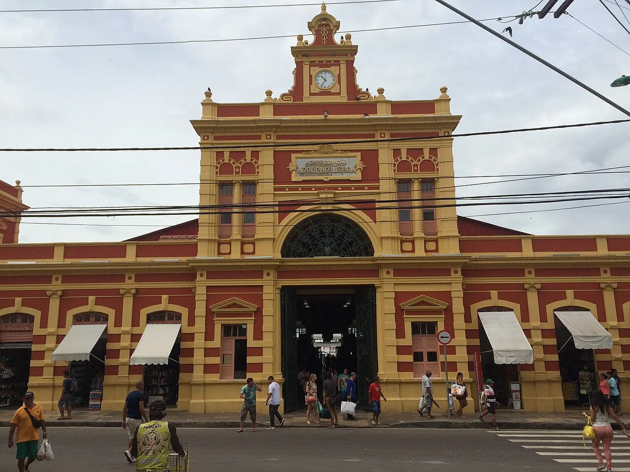 Fachada de hierro roja y amarilla del Mercado Municipal Adolpho Lisboa, con torre del reloj, arco de entrada y personas caminando en la acera bajo un cielo nublado.