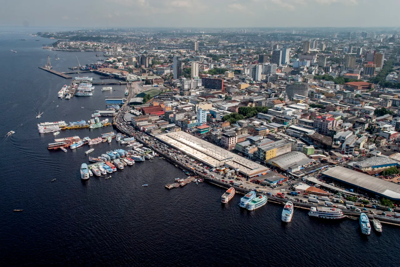 Vista aérea do porto de Manaus, com embarcações atracadas ao longo dos cais flutuantes e os prédios do centro ao fundo.