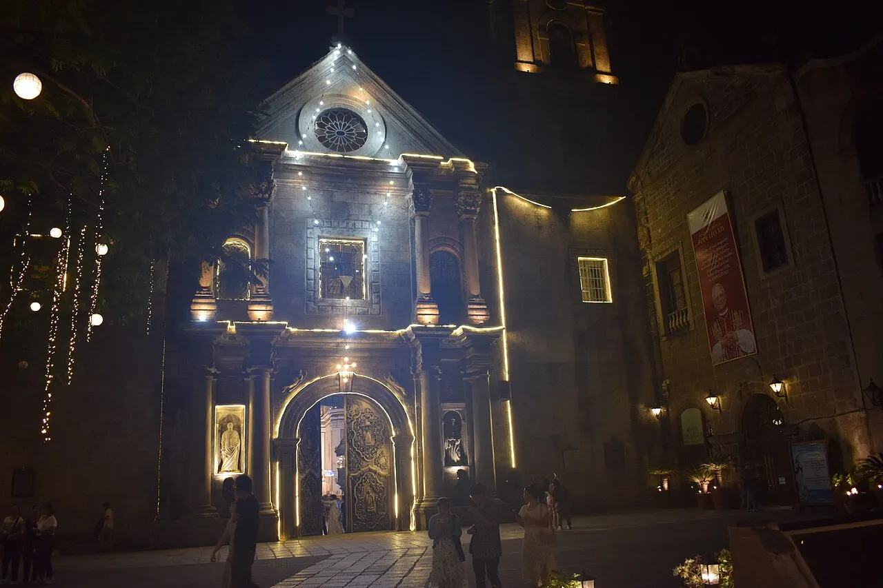 Fachada de igreja de pedra iluminada à noite, com um frontão triangular sobre a porta principal.