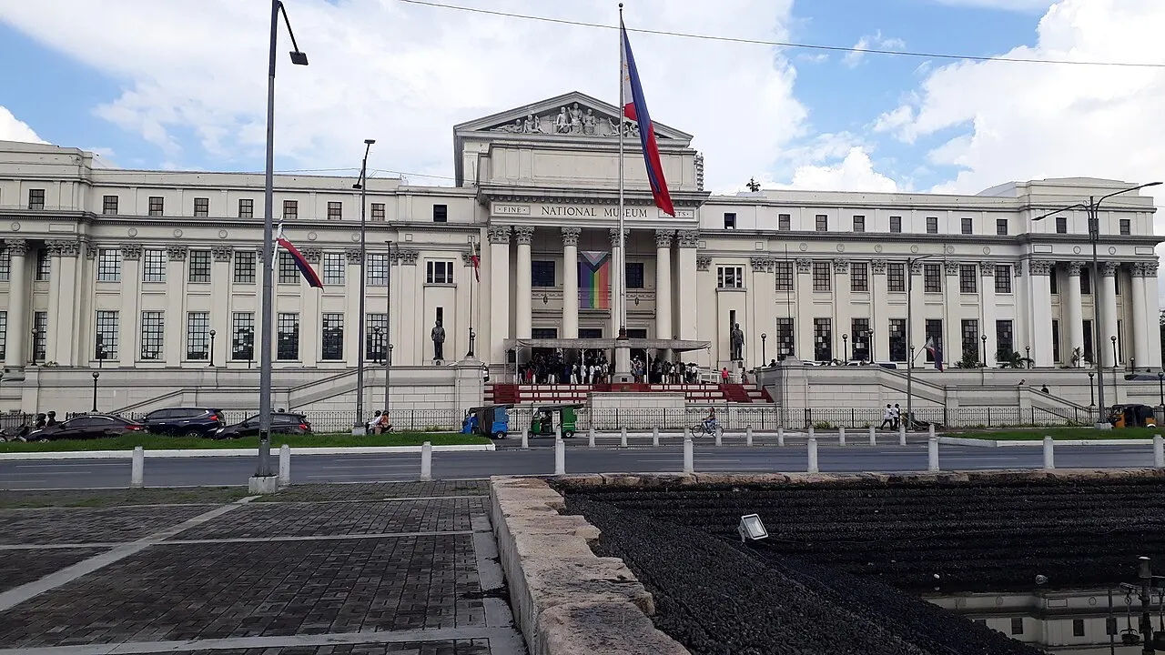Fachada neoclássica do Museu Nacional de Belas Artes em Manila, com colunas, frontão triangular e bandeiras hasteadas em frente, sob céu com nuvens.
