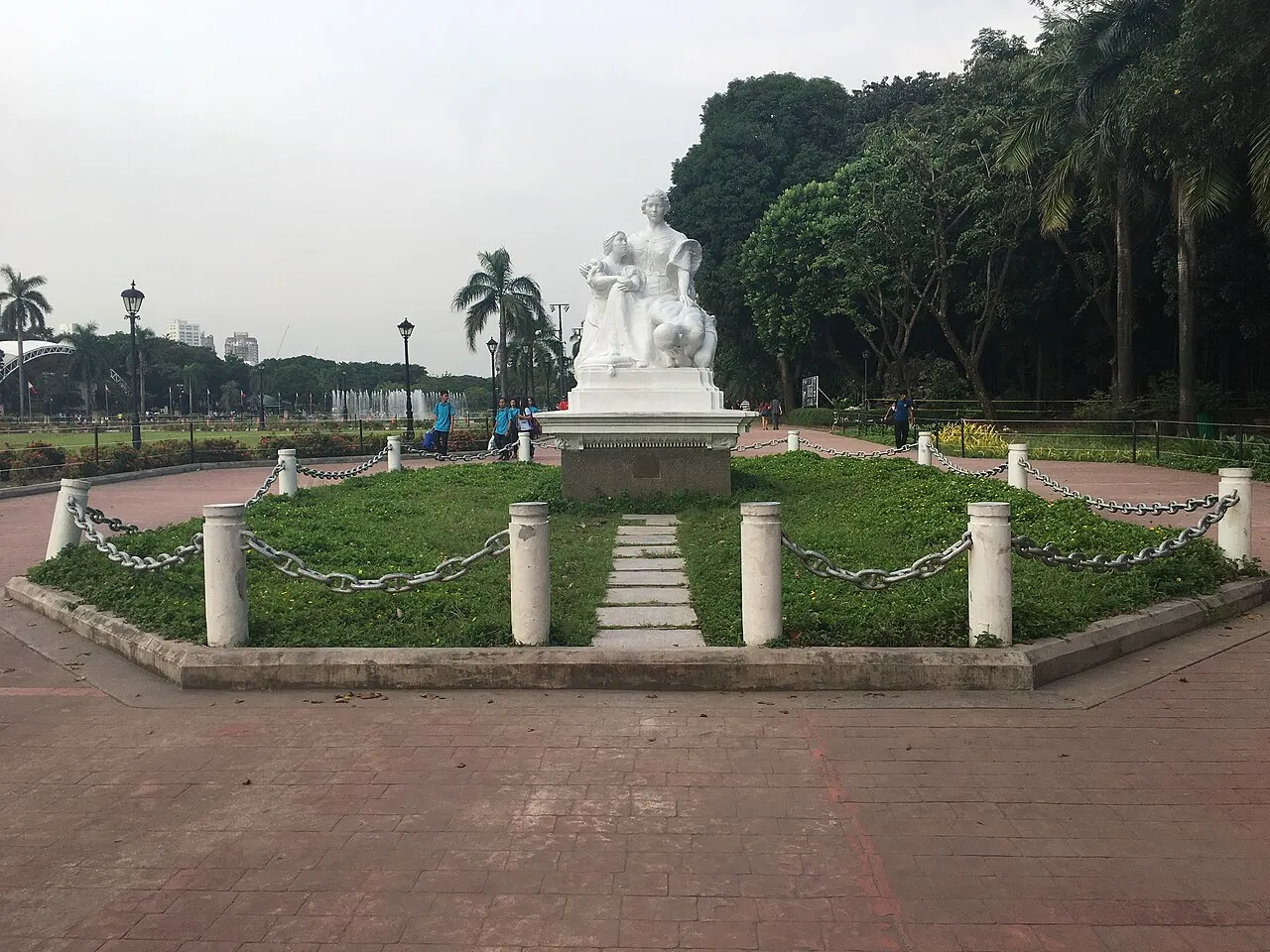 Estátua branca de uma mulher sentada com crianças, sobre um pedestal cercado de grama.