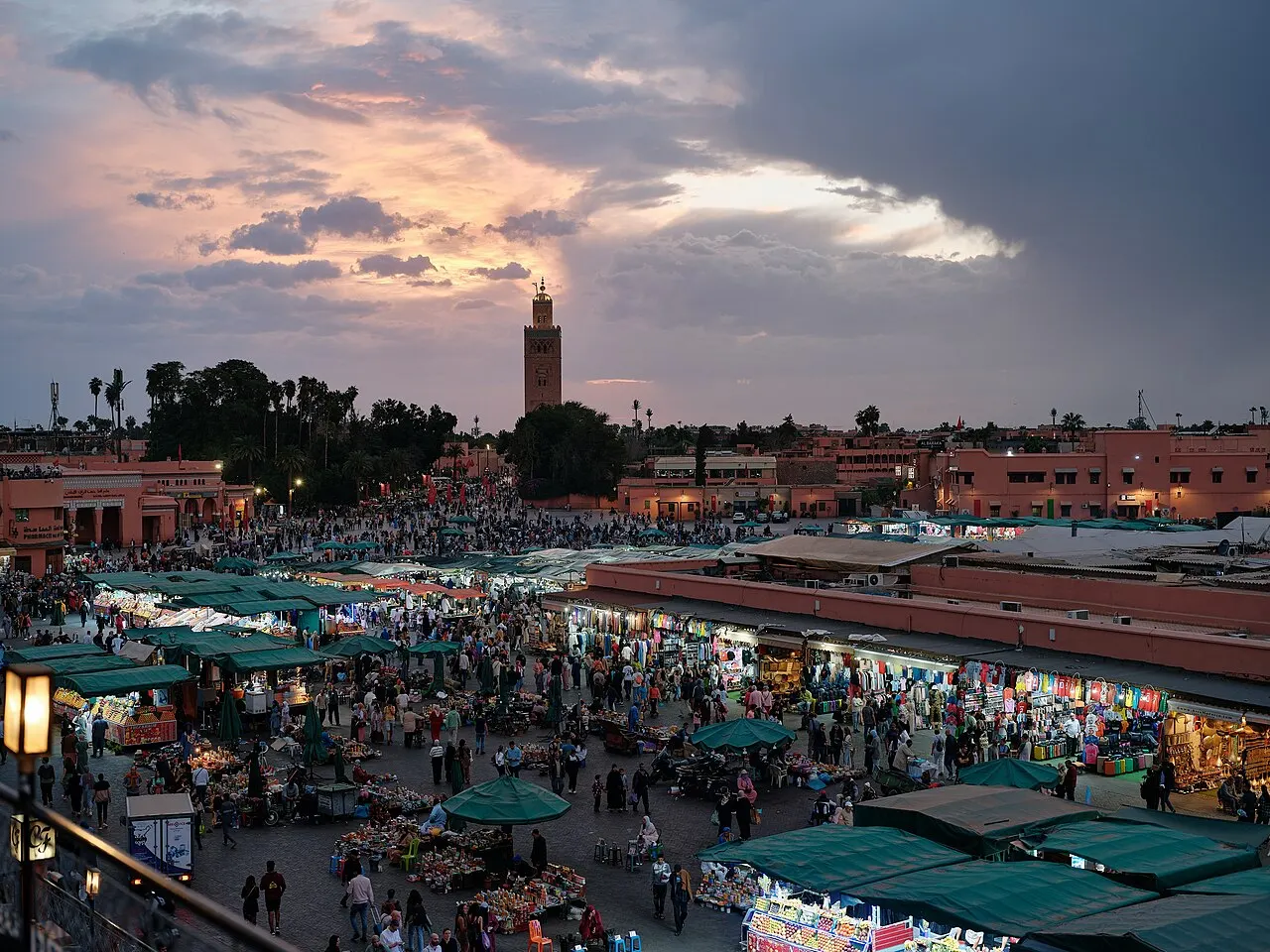 Vista aérea da praça Jemaa el-Fna cheia de barracas e gente ao entardecer, com um minarete ao fundo sob céu de nuvens rosadas e douradas.