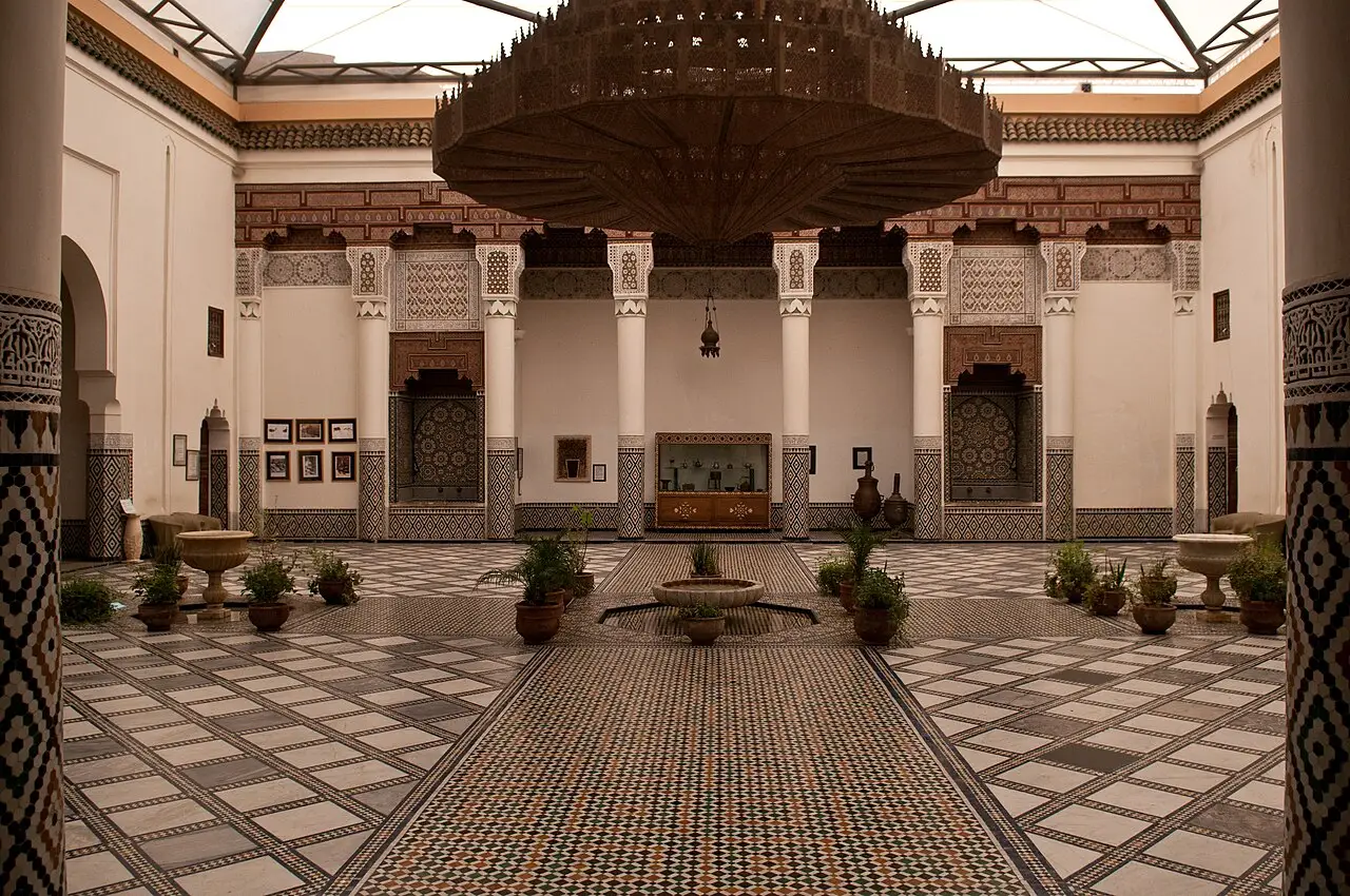 Pátio do Museu de Marrakech, com um grande lustre de metal, colunas brancas, piso geométrico e vasos em volta da fonte.