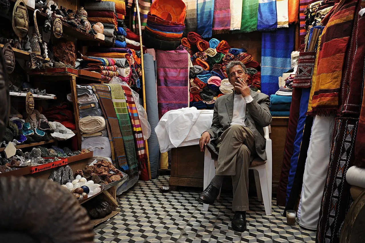 Um homem sentado numa loja dos souks, cercado de tecidos coloridos dobrados, máscaras de madeira e pedras à venda, num piso de ladrilhos xadrez.