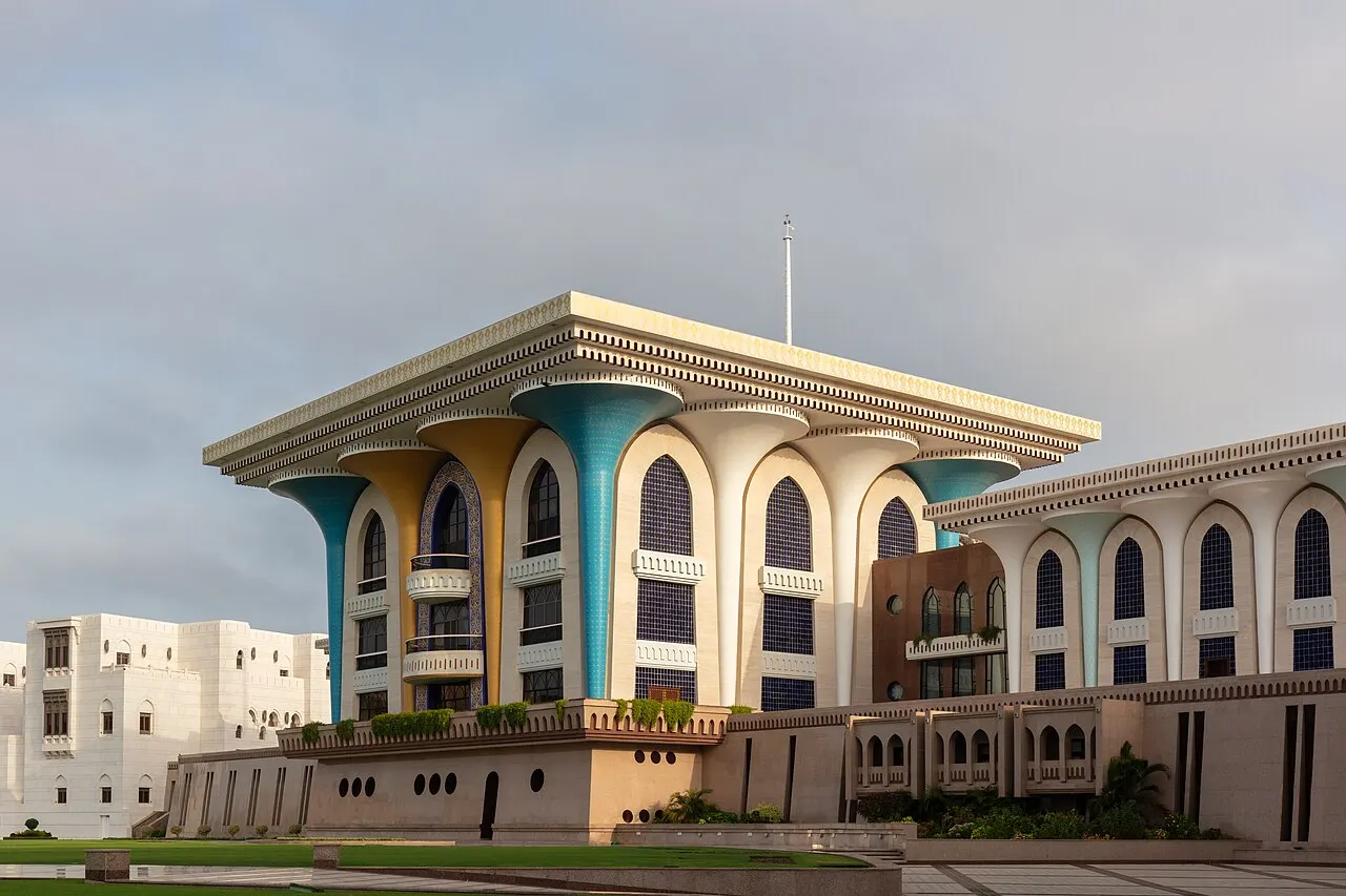 A palace with a broad flat roof held up by cup-shaped columns painted blue and gold, with white buildings beside it and a cloudy sky.