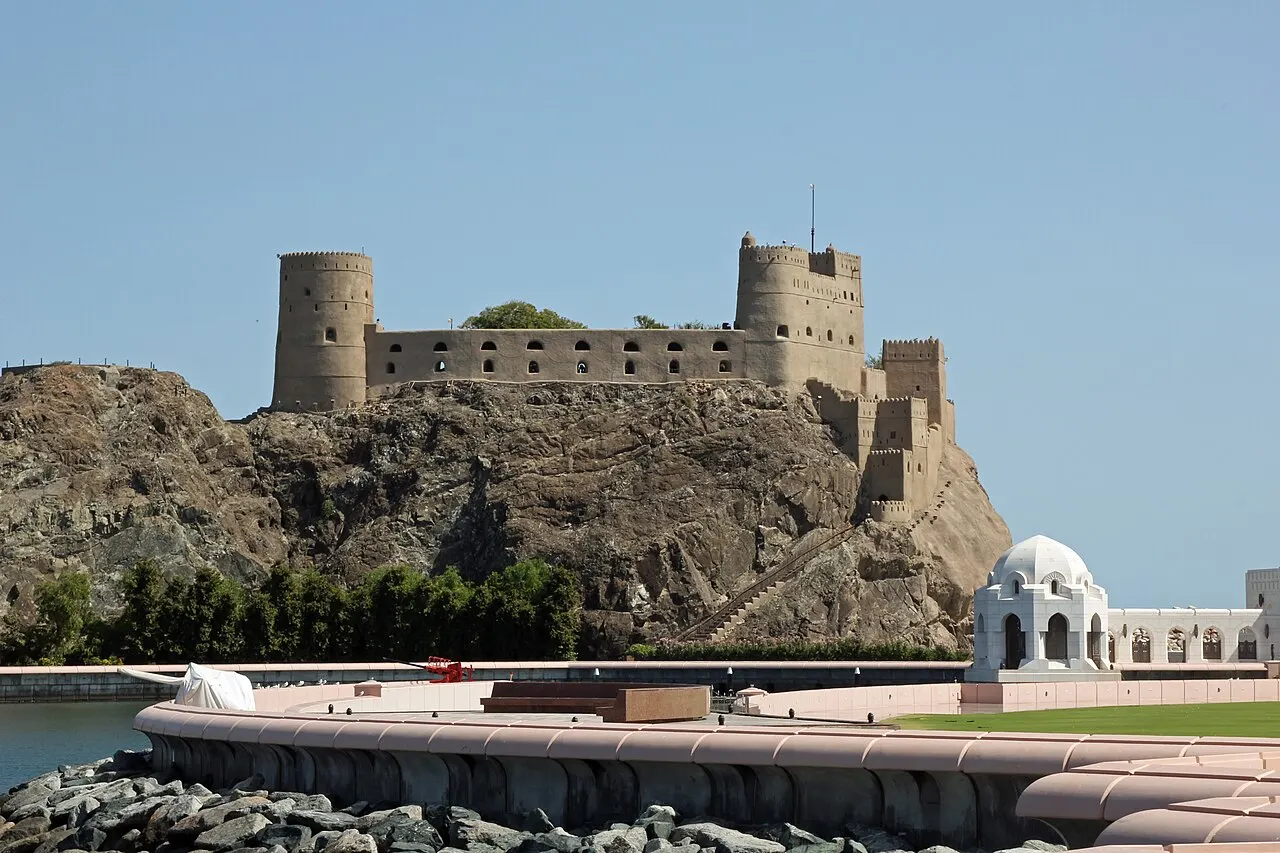A stone fort with two round towers on top of a dark rock, above a curved quay, with a white domed pavilion on the right.