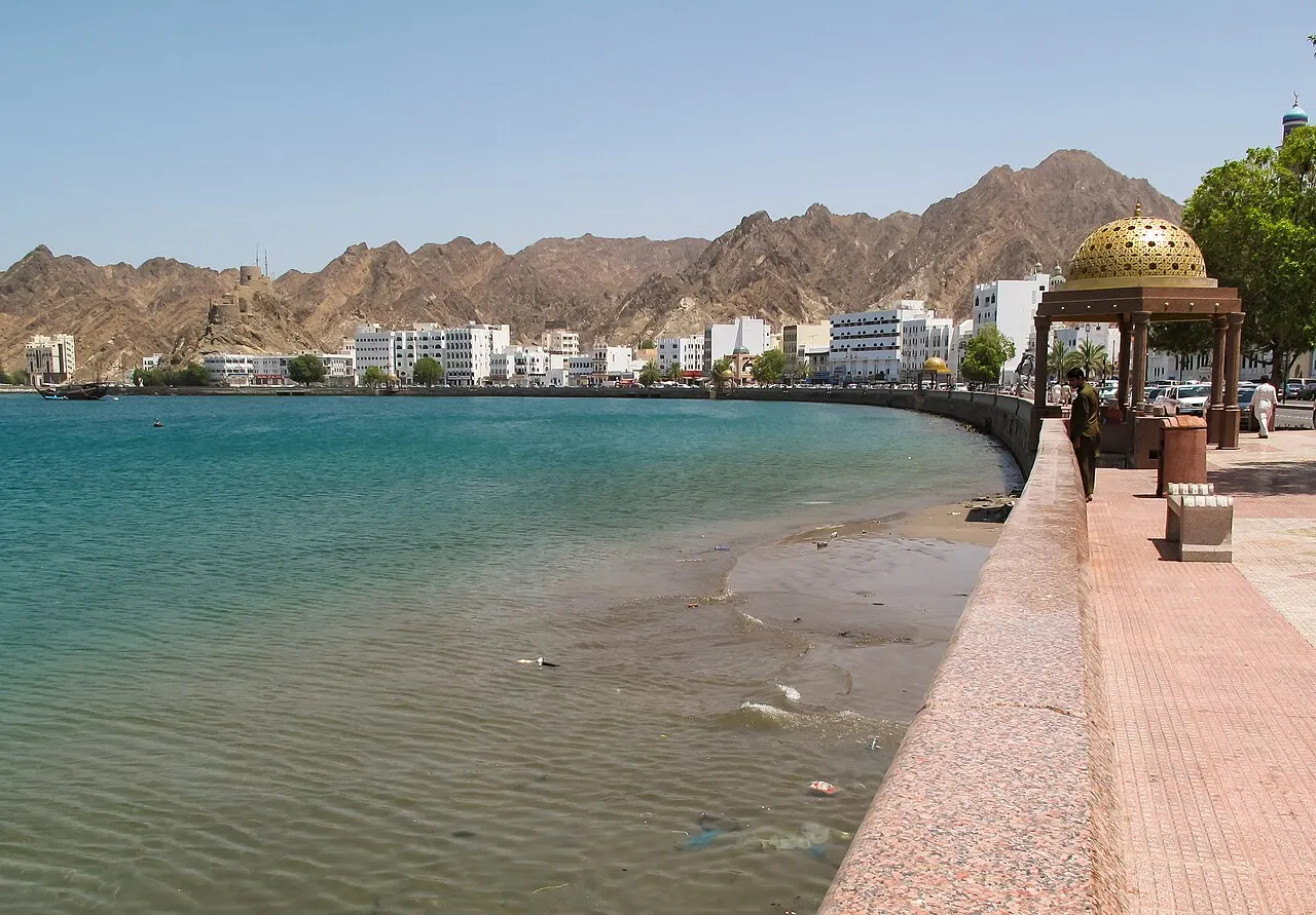 Un paseo de suelo rosado junto a agua verde y poco profunda, con un quiosco de cúpula dorada, edificios blancos en la curva de la bahía y montañas áridas al fondo.