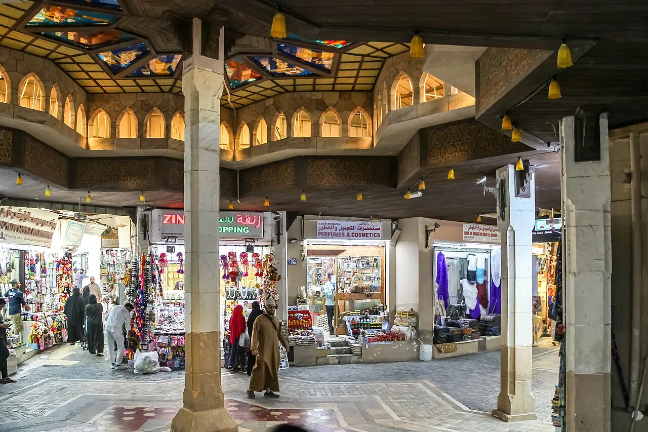 Un patio cubierto con columnas de piedra y faroles colgantes, rodeado de tiendas iluminadas de telas y recuerdos, con gente caminando al fondo.
